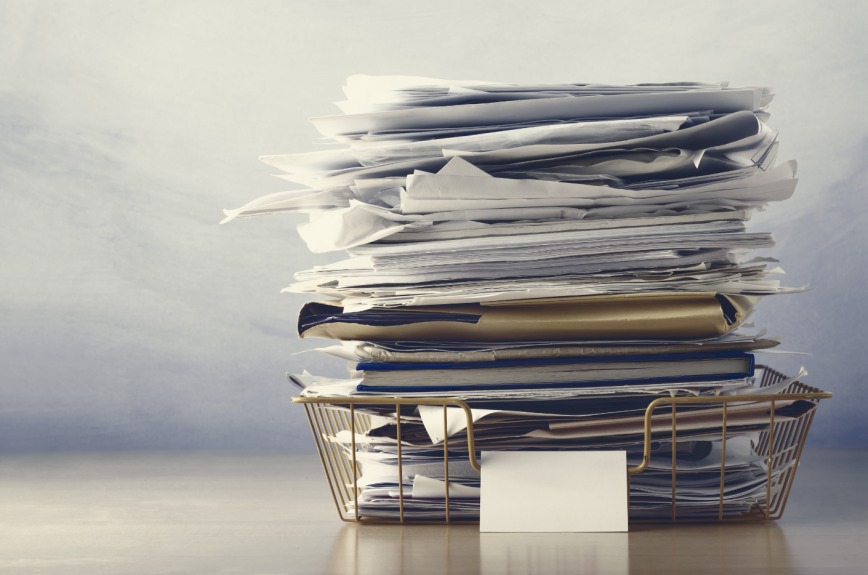 Tall stack of scattered papers and folders in a metal filing basket on a desk.
