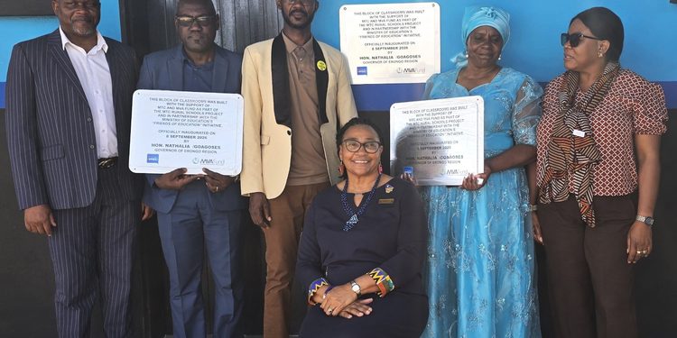 Group of six adults standing with plaques in front of a bright blue wall; woman sits front center in a dark dress with colorful accents.