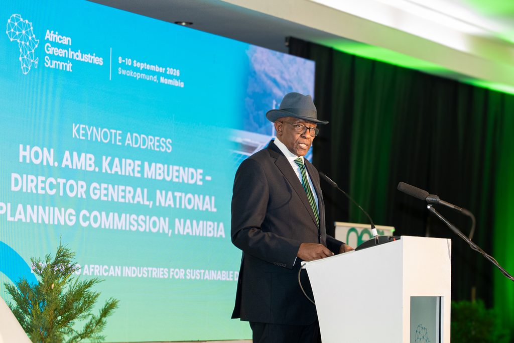 Speaker at a podium delivering the keynote at the African Green Industries Summit, with a blue screen behind him displaying event details