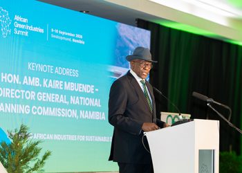 KAIRE MBUENDE6 - The Brief | Namibia's Leading Business & Financial News Speaker at a podium delivering the keynote at the African Green Industries Summit, with a blue screen behind him displaying event details