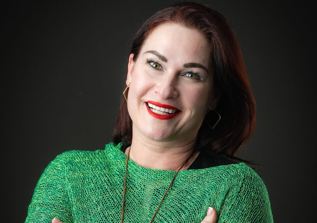 Portrait of a smiling woman with red lipstick wearing a green knit sweater against a dark background.