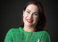 Portrait of a smiling woman with red lipstick wearing a green knit sweater against a dark background.