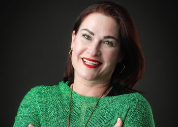 Natasja Beyleveld1 - The Brief | Namibia's Leading Business & Financial News Portrait of a smiling woman with red lipstick wearing a green knit sweater against a dark background.