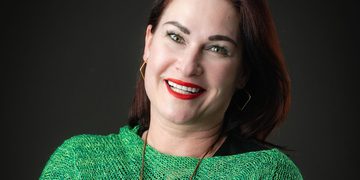 Portrait of a smiling woman with red lipstick wearing a green knit sweater against a dark background.