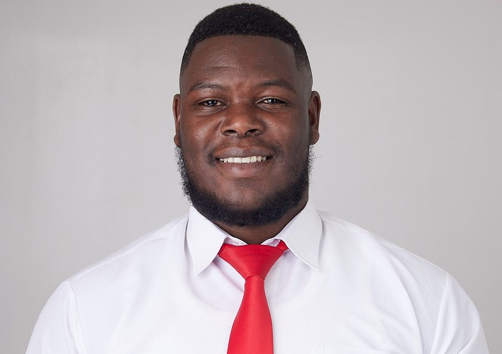 Smiling man in a white dress shirt and bright red tie, facing the camera against a light gray background.
