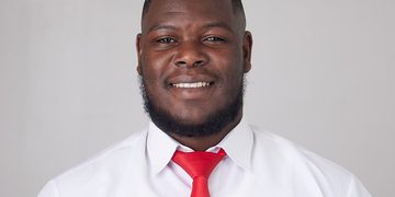 Smiling man in a white dress shirt and bright red tie, facing the camera against a light gray background.