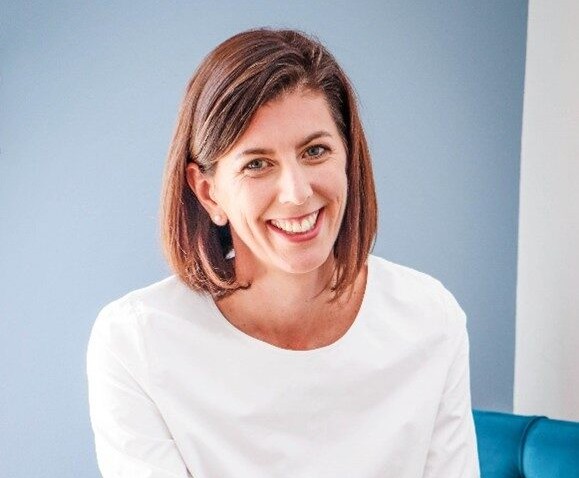 Smiling woman with shoulder-length brown hair in a white blouse sits against a blue wall, looking at the camera.