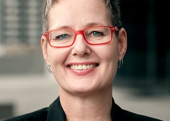 Eline-Van-Der-Linden - The Brief | Namibia's Leading Business & Financial News Smiling woman with short gray hair and red rectangular glasses, wearing a black blazer, outdoors near a modern building grounds.