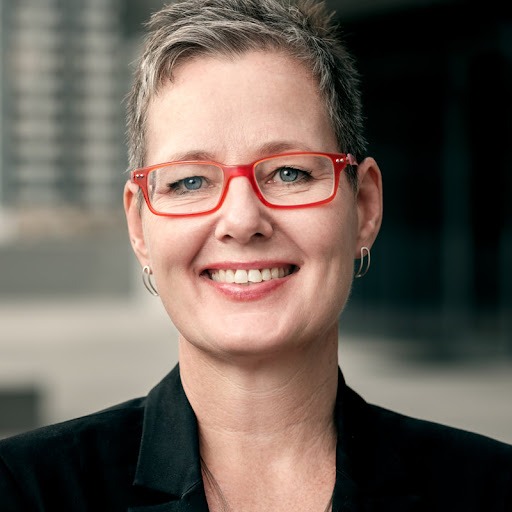 Eline-Van-Der-Linden - The Brief | Namibia's Leading Business & Financial News Smiling woman with short gray hair and red rectangular glasses, wearing a black blazer, outdoors near a modern building grounds.