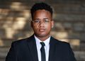 Arney Tjaronda - The Brief | Namibia's Leading Business & Financial News Professional portrait of a man in a black suit and tie wearing clear-framed glasses, looking at the camera against a blurred brick wall background.