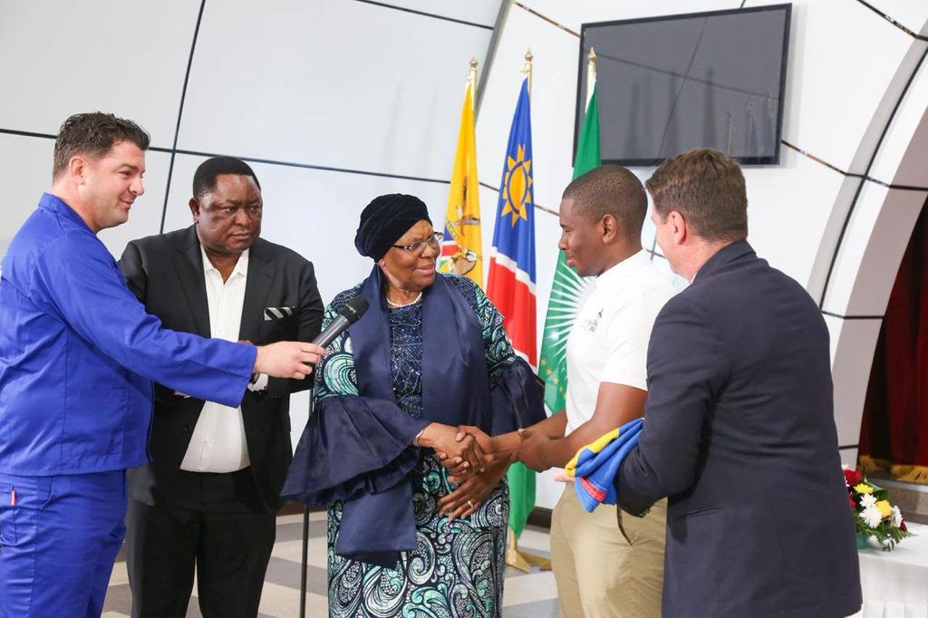 WhatsApp Image 2026-09-14 at 114824 AM - The Brief | Namibia's Leading Business & Financial News Interviewer in a blue suit hands a microphone to a woman in a patterned dress as she shakes hands with a young man; flags behind them.