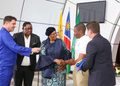 Interviewer in a blue suit hands a microphone to a woman in a patterned dress as she shakes hands with a young man; flags behind them.