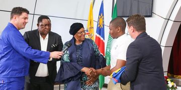 Interviewer in a blue suit hands a microphone to a woman in a patterned dress as she shakes hands with a young man; flags behind them.