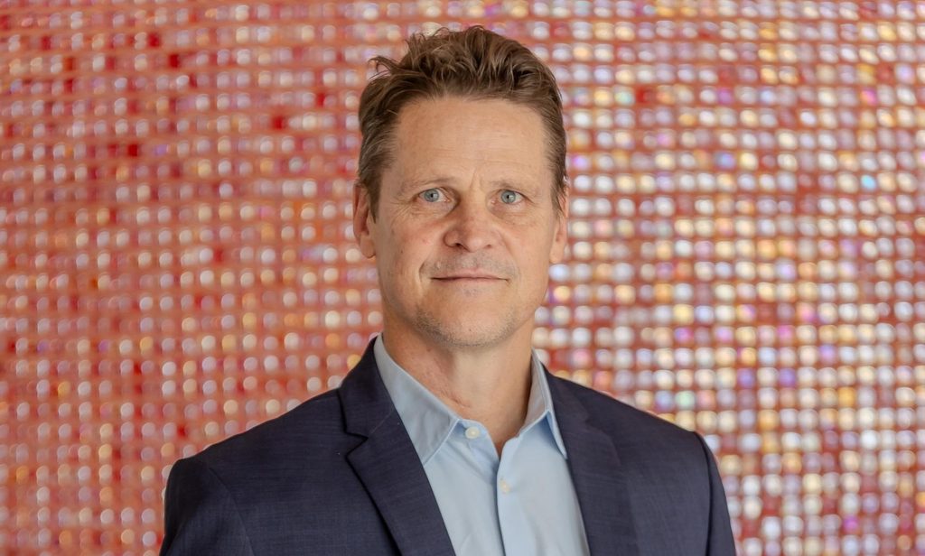 Riaan Vermeulen - The Brief | Namibia's Leading Business & Financial News Portrait of a man in a navy blazer and light blue shirt, standing in front of a colorful tiled background.