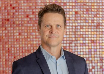 Portrait of a man in a navy blazer and light blue shirt, standing in front of a colorful tiled background.