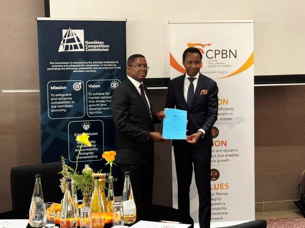 WhatsApp Image 2026-09-15 at 104858 AM - The Brief | Namibia's Leading Business & Financial News Two men in suits shake hands and exchange a blue document at a formal event, with Namibian Competition Commission and CPBN banners in the background.