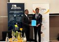 Two men in suits shake hands and exchange a blue document at a formal event, with Namibian Competition Commission and CPBN banners in the background.