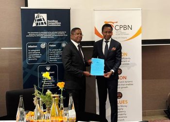 Two men in suits shake hands and exchange a blue document at a formal event, with Namibian Competition Commission and CPBN banners in the background.