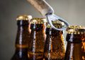 Beer bottles with caps being pried open by a metal bottle opener, close-up shot of condensation-streaked bottles in a row.