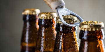 Beer bottles with caps being pried open by a metal bottle opener, close-up shot of condensation-streaked bottles in a row.