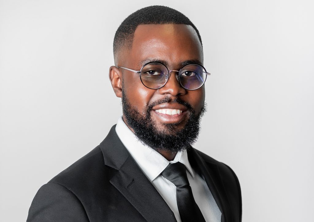 Evoto - The Brief | Namibia's Leading Business & Financial News Smiling man wearing a black suit, white shirt, black tie, and round glasses against a light gray background.