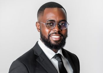 Evoto - The Brief | Namibia's Leading Business & Financial News Smiling man wearing a black suit, white shirt, black tie, and round glasses against a light gray background.
