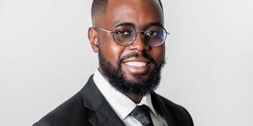 Smiling man wearing a black suit, white shirt, black tie, and round glasses against a light gray background.