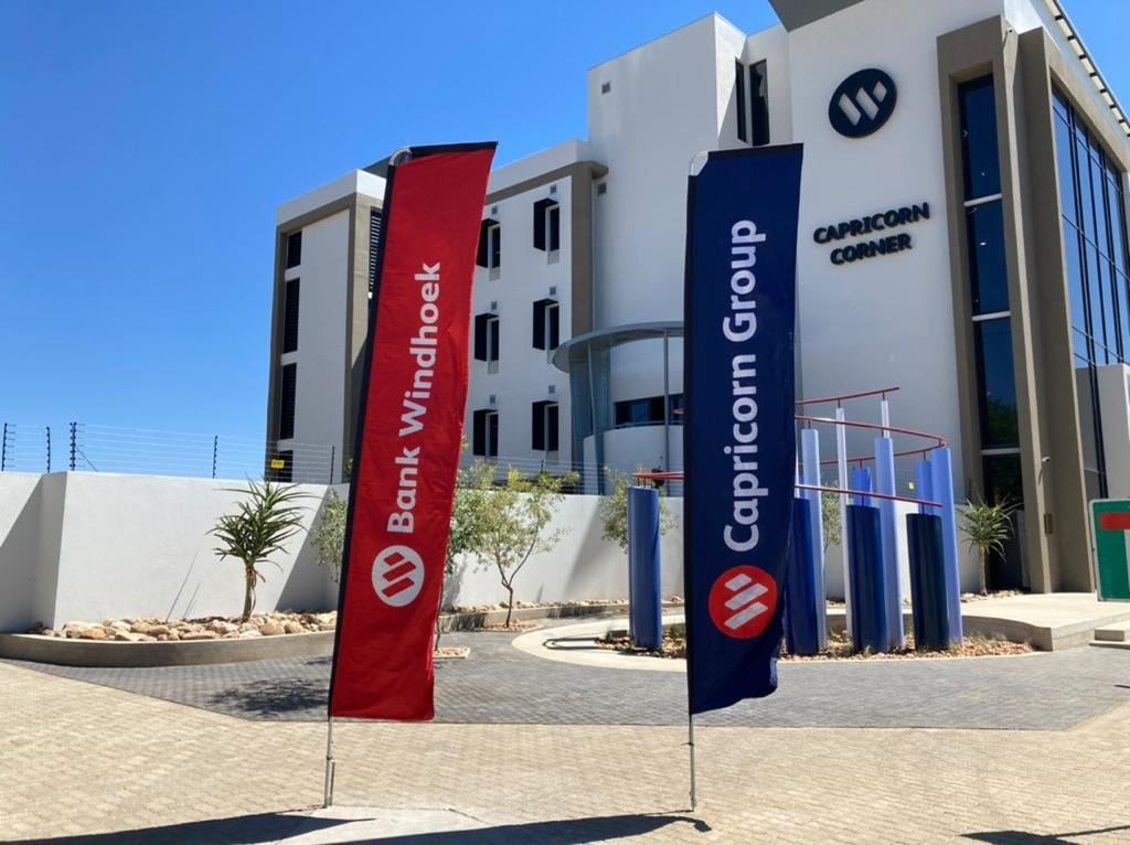 Modern corporate building with red Bank Windhoek and blue Capricorn Group flags in front of a clear sky.