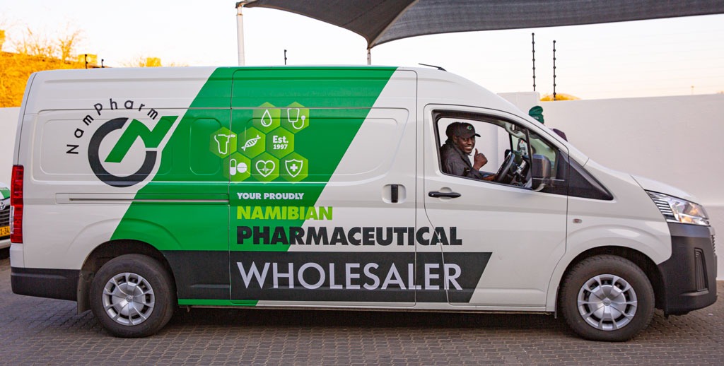 White promotional van for a Namibian pharmaceutical wholesaler with green branding and a driver waving from the cab window.