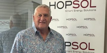 Older man with gray hair in a patterned shirt stands in front of a HopSOL banner reading 'Smart Energy Solutions'.