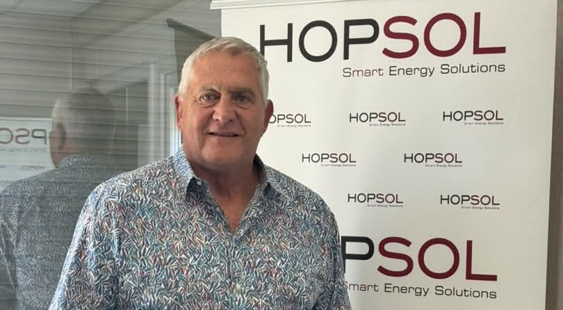 Older man with gray hair in a patterned shirt stands in front of a HopSOL banner reading 'Smart Energy Solutions'.