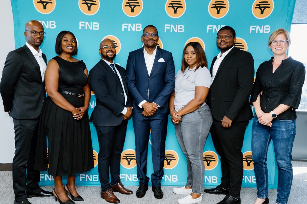 WhatsApp Image 2026-09-20 at 24914 PM - The Brief | Namibia's Leading Business & Financial News Seven professionals posing for a group photo in front of a teal FNB banner, smiling and dressed in business attire.