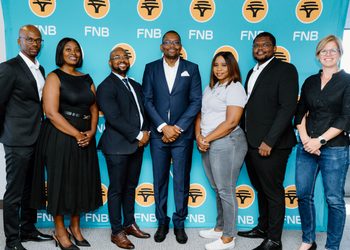 WhatsApp Image 2026-09-20 at 24914 PM - The Brief | Namibia's Leading Business & Financial News Seven professionals posing for a group photo in front of a teal FNB banner, smiling and dressed in business attire.