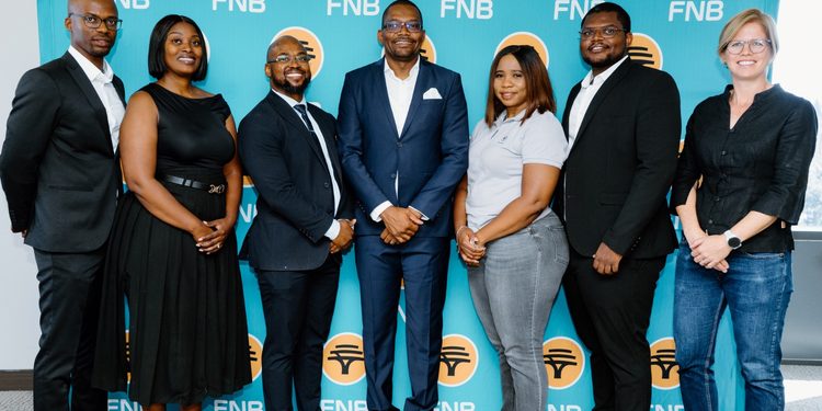 Seven professionals posing for a group photo in front of a teal FNB banner, smiling and dressed in business attire.