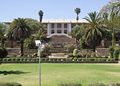 Beige, two-story building perched above terraced gardens with palm trees and wide stone steps, set in a lush green lawn.