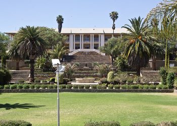 PARLIAMENT - The Brief | Namibia's Leading Business & Financial News Beige, two-story building perched above terraced gardens with palm trees and wide stone steps, set in a lush green lawn.