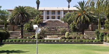 Beige, two-story building perched above terraced gardens with palm trees and wide stone steps, set in a lush green lawn.