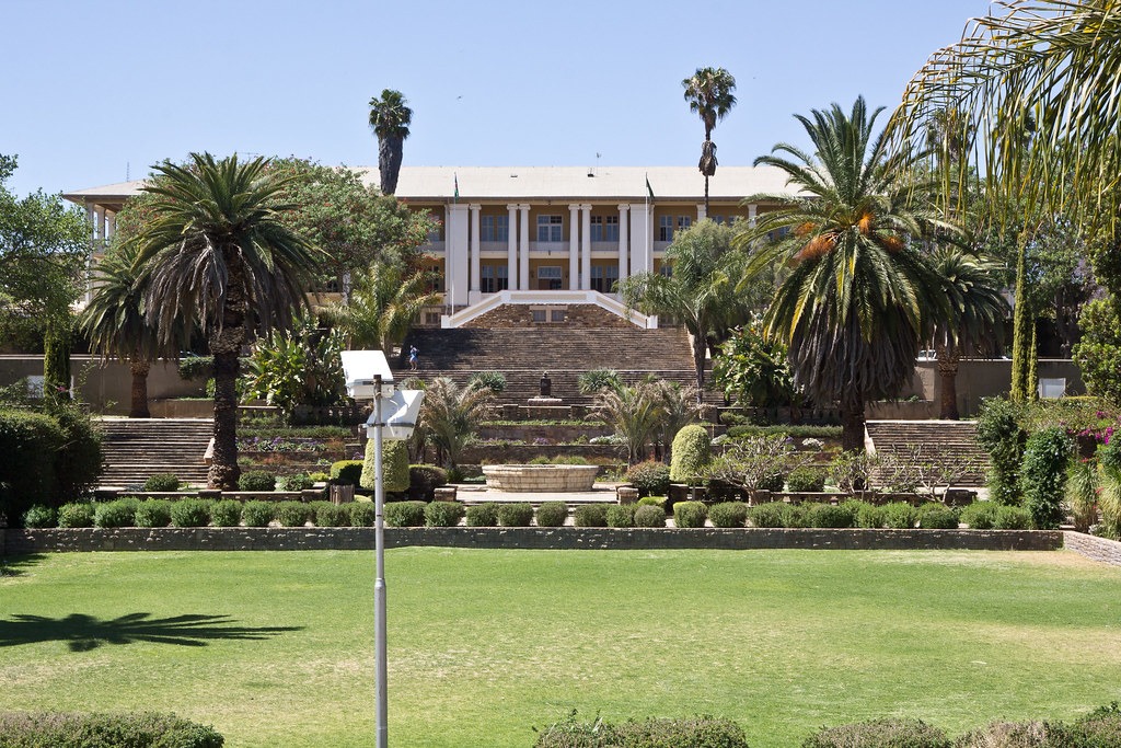 PARLIAMENT - The Brief | Namibia's Leading Business & Financial News Beige, two-story building perched above terraced gardens with palm trees and wide stone steps, set in a lush green lawn.