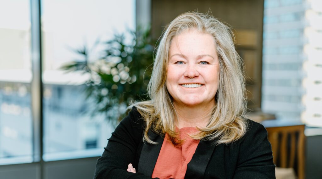 Angelique Peake - The Brief | Namibia's Leading Business & Financial News Smiling woman with blonde hair, coral blouse, and black blazer in an office setting.