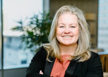 Angelique Peake - The Brief | Namibia's Leading Business & Financial News Smiling woman with blonde hair, coral blouse, and black blazer in an office setting.