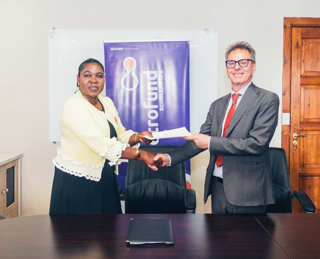 PETROFUND and Saipem MoU Signing Ceremony 2jpg - The Brief | Namibia's Leading Business & Financial News Two professionals shake hands while exchanging a document in an office, with a purple banner reading 'Fund Scholarships' in the background.