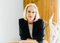 Professional woman with blonde hair in a black blazer, seated indoors beside a window. (informative)