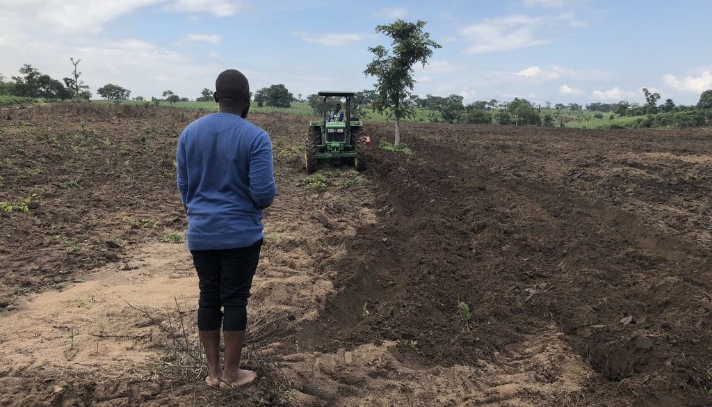Man in a blue shirt standing barefoot as a green tractor digs a long trench in a tilled field under a blue sky.