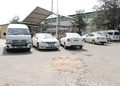 White van and four white cars parked in a gravel lot under a metal canopy beside a warehouse/storage building.
