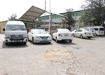 GOVERNMENT CARS - The Brief | Namibia's Leading Business & Financial News White van and four white cars parked in a gravel lot under a metal canopy beside a warehouse/storage building.