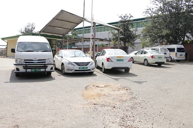 GOVERNMENT CARS - The Brief | Namibia's Leading Business & Financial News White van and four white cars parked in a gravel lot under a metal canopy beside a warehouse/storage building.