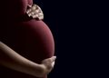 Pregnant person cradling their belly with both hands against a dark background.