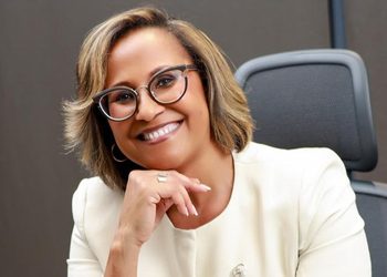 Smiling woman with short hair and round glasses seated in an office setting, resting her chin on her hand.