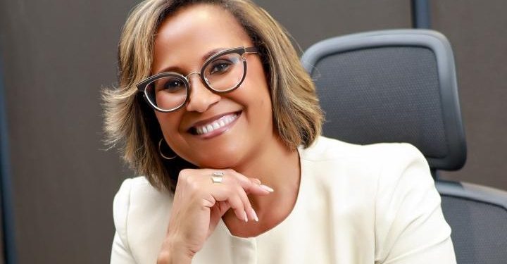 Smiling woman with short hair and round glasses seated in an office setting, resting her chin on her hand.