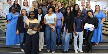 Diverse group of professionals posing on stairs in a bright lobby, some holding folders or boxes and smiling at the camera.